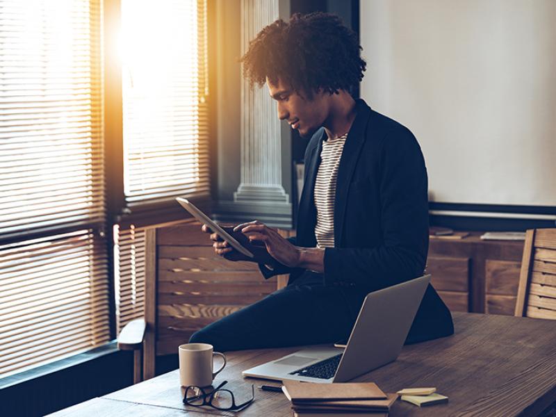 Man sitting on table and working on tablet, on table a laptop and notebooks are placed